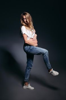 Front view of pretty and fashionable pregnant woman wearing in jeans and white t shirt embracing stomach. Future mother smiling at camera,holding hands on her naked tummy. Concept of pregnant fashion.