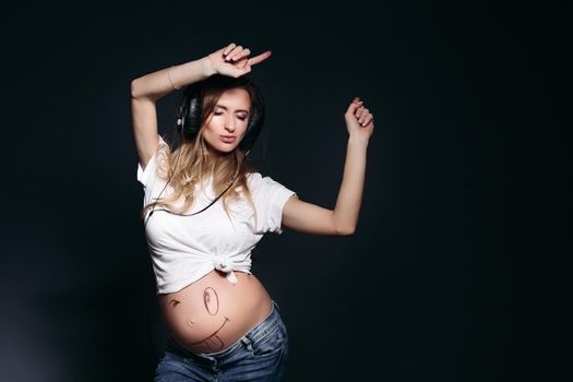 Happy and young pregnant mom dancing, smiling at camera and rising up by hands. Beautiful expectant mother listening music by big earphones and holding arms up. Female expacting her child.