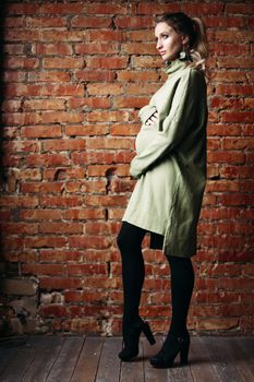 Portrait of happy pregnant brunette girl posing against brick wall. Stylish woman wearing in khaki dress, holding pregnant belly by hands, looking at side. Concept of pregnancy and paternity.