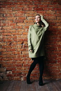 Portrait of happy pregnant brunette girl posing against brick wall. Stylish woman wearing in khaki dress, holding pregnant belly by hands, looking at side. Concept of pregnancy and paternity.