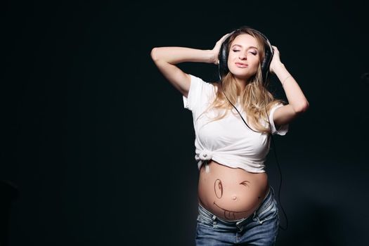 Portrait of beautiful pregnant woman with long hair dancing, smiling with closed eyes. Expectant mother with naked tummy listening music and holding big earphones by hands. Stylish mom wearing in jeans.