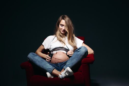 Front view of adorable expectant mother sitiing on red chair and holding earphones on her tummy with painted smile. Lady wearing blue jeans and white tee shirt. Concept of maternity. Black background.
