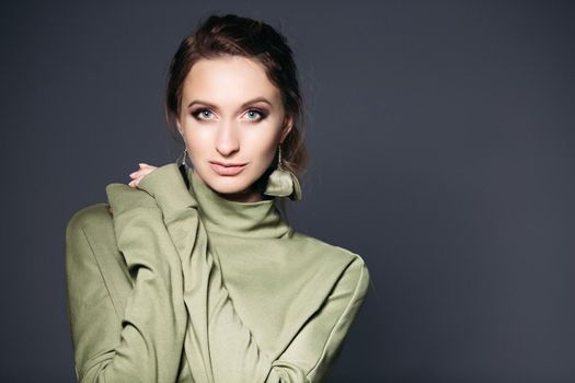 Beauty portrait in dark studio of brunette woman with wearing in khaki dress, big blue eyes, plump lips, posing. Gorgeous female with perfect make up holding hands on shoulder, looking at camera.