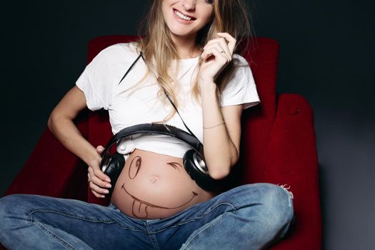 Crop of young pregnant mother sitiing on red chair smiling and holding earphones on her tummy with painted smile. Pretty mom wearing blue jeans and white t-shirt. Black studio background.
