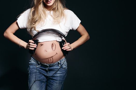 Happy and smiling pregnant female wearing white t shirt and jeans with big earphones on tummy. Expectant mother painting funny smile on her naked belly. Concept of maternety and health care.