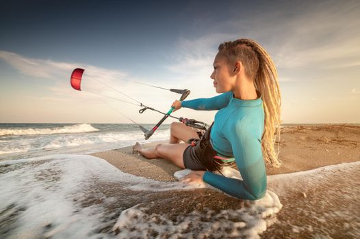 Attractive caucasian woman kitesurfer in a neoprene wetsuit and with dreadlocks on her head is resting lying on a sandy beach on the shore holding her kite in the wind.