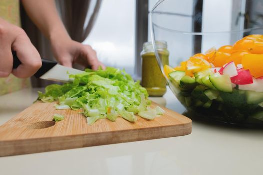 Female hands cut lettuce leaves with a knife against the background of a bowl with chopped vegetables. Vegetarian food preparation.