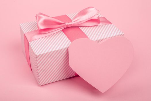 Valentine Day gift in a box wrapped in striped paper and tied with silk ribbon bow and heart shapes greeting card on pink background with copy space for text