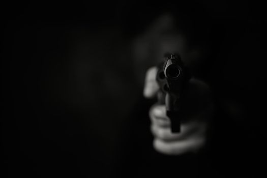 Threat of firearms. Muzzle of pistol in person's hand is pointed at camera. Man criminal holds revolver on dark background. Attack or defense. Black and white photo.