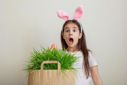A surprised girl in pink bunny ears hold pink egg from a paper bag with grass.Copy space