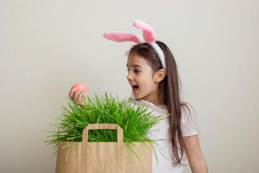A beautiful little surprised girl in pink bunny ears hold pink egg from a paper bag with grass.Copy space