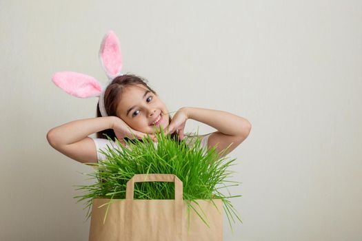 A portrait of a charming girl with bunny ears stands next to a paper bag with juicy green grass. Copy space.