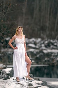 A young woman in a white dress on the shore of a lake in the winter in a snowy landscape.