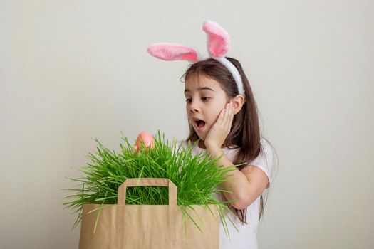 A little surprised girl in a white T-shirt and pink bunny ears took out a pink egg from a paper bag with grass.Copy space