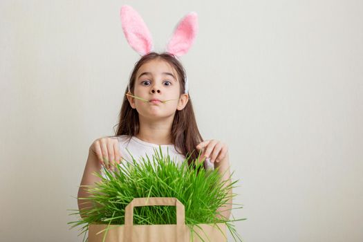 A portrait of a funny girl with bunny ears stands next to a paper bag with juicy green grass. Copy space.
