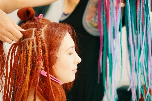 Barber braids dreadlocks. Hairdresser weaves braids with kanekalon to young girl head, making creative hairstyle with thick plaits. Cool hairstyle made of artificial hair. Rainbow Afro-pigtails.