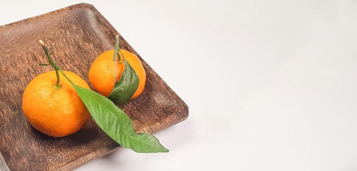 Two fresh ripe tangerines with leaves and branches on a dark square wooden plate, close-up, space for text on the right.