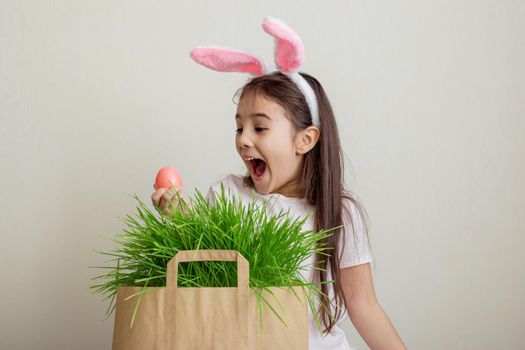 A funny little surprised girl in pink bunny ears took out a pink egg from a paper bag with grass.Copy space