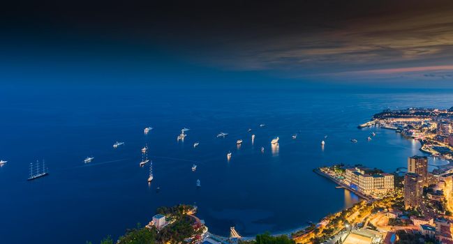 A lot of yachts and motor boats with night illumination are moored in mediterranean sea, aerial view of bay of Monaco - Monte-Carlo at dusk, cityscape with night illumination. High quality photo
