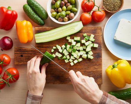 Step by step recipe for salad horiatiki, wooden board for cutting vegetables and ingredients. Hands with knife, chopping cucumber