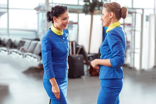 Two joyful women stewardesses in aviation uniform chatting and smiling while waiting for the plane at airport