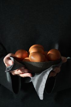 Unusual Easter on a dark background. A bowl of brown eggs with hands. Darkness, rays of sunlight shine on the eggs. The concept of a new life, rebirth. Rustic style. Vertical, copy space