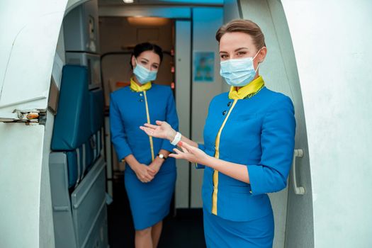 Two women stewardesses wearing protective face masks while standing near airplane door and welcoming passengers