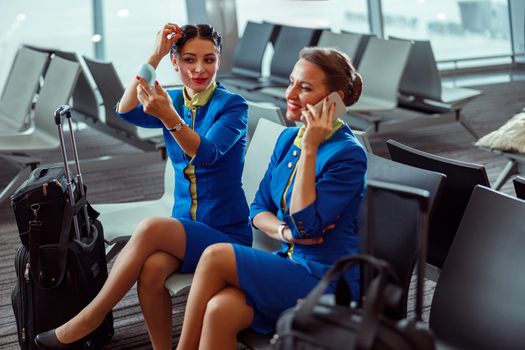 Female flight attendant looking in travel mirror and smiling while colleague talking on cellphone in departure lounge