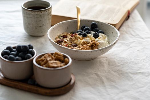 Reading book and eating healthy lifestyle breakfast with granola muesli and yogurt in bowl on white table background, cereal grain food with nuts seed. Organic morning diet meal with oat