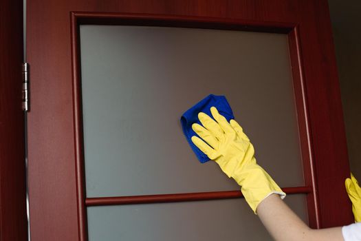 Crop of housewife hands wearing yellow protective gloves, cleaning dirty window in doors. Housemaid wiping glass with rag. Concept of housework and service.
