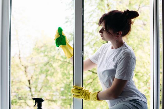 Housemaid wearing white t shirt and yellow protective gloves wiping glass with rag. View of brunette housewife cleaning big dirty window. Concept of housework and apartment service.