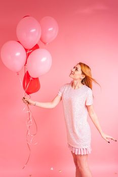 Beautiful redhead girl with red heart baloon posing. Happy Valentine's Day concept. Studio photo of beautiful ginger girl dancing on pink background.