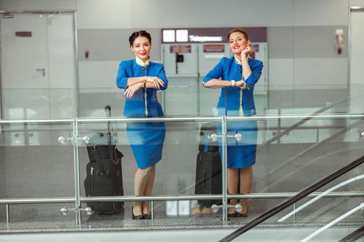 Cheerful female flight attendants in air hostess uniform looking at camera and smiling while standing near trolley luggage bags