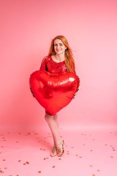 Beautiful redhead girl with red heart baloon posing. Happy Valentine's Day concept. Studio photo of beautiful ginger girl dancing on pink background.