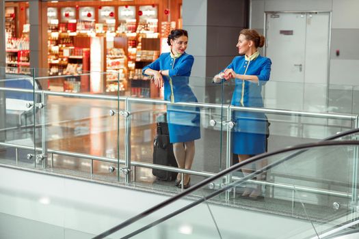Women stewardesses in aviation air hostess uniform talking and smiling while standing near travel bags in passenger departure terminal