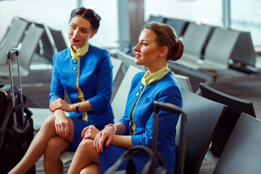 Smiling women stewardesses in air hostess uniform sitting on chairs in passenger departure lounge