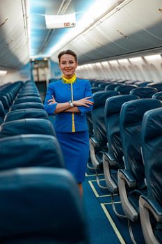 Joyful flight attendant in air hostess uniform keeping arms crossed and smiling while standing in aisle of airplane salon
