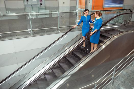 Smiling women flight attendants with travel bags wearing aviation air hostess uniform while standing on escalator stairs