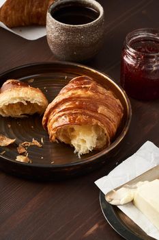 Morning breakfast with croissant on plate, cup of coffee, jam and butter. Dark brown table , morning routine, slow life. Vertical, side view