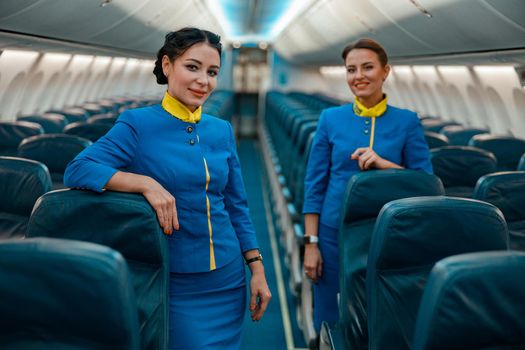 Joyful women flight attendants in air hostess uniform looking at camera and smiling while standing near passenger seats inside airplane