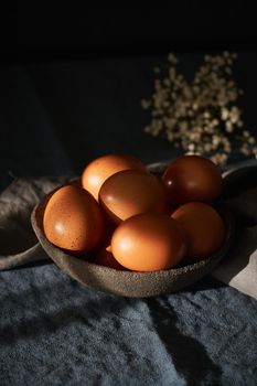 Unusual Easter on dark background. Bowl of brown eggs on dark blue table, flowers. Darkness, rays of sunlight shine on eggs. Concept of new life, rebirth. Side view, copy space, vertical