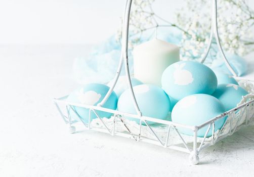 Easter. Holiday. Light white background, gentle pastel colors. Blue eggs with image of rabbit in basket. Flowers in background, side view, copy space