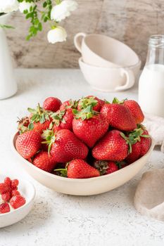 Side view of bowl with strawberry on white table, bottle with milk, flower. Still-life photo with summer berries, vertical