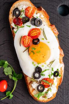 Toasted bread toast with fried eggs with yellow yolk and tomatoes, olives, sprinkled with herbs on dark wooden serving board on blue denim napkin, vertical, top view, close up