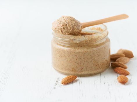 Almond butter, raw food paste made from grinding almonds into nut butter, crunchy and stir, white wooden table, glass jar, side view, close up, copy space