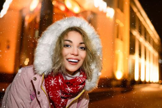 portrait of a charming woman on the snowy street, in a winter jacket, smiles happily around the evening lights of the city, she is shines with happiness