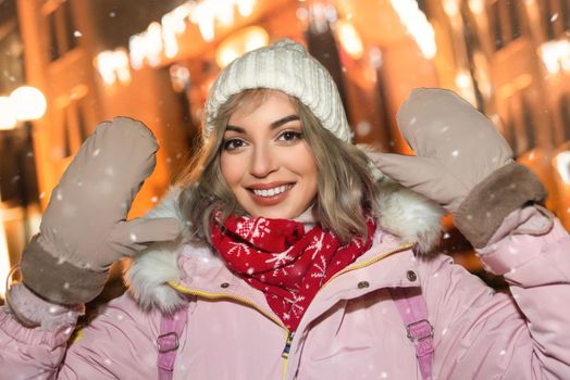 portrait of a charming woman on a snow-covered street, wearing a winter jacket, smiling happily against the city's evening lights, she looks into the camera and raises her hands up