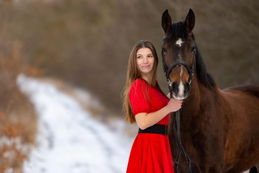 Portrait of a beautiful girl in a red dress standing with a horse on the background of a winter road a