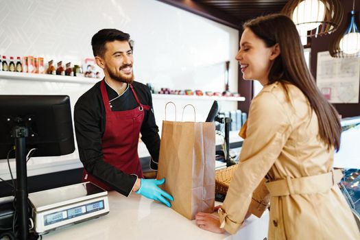 Shop assistant handling shopping bag to female customer in grocery store, close up