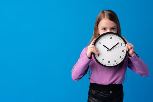 Beautiful teen girl holding wall clock over blue background, close up